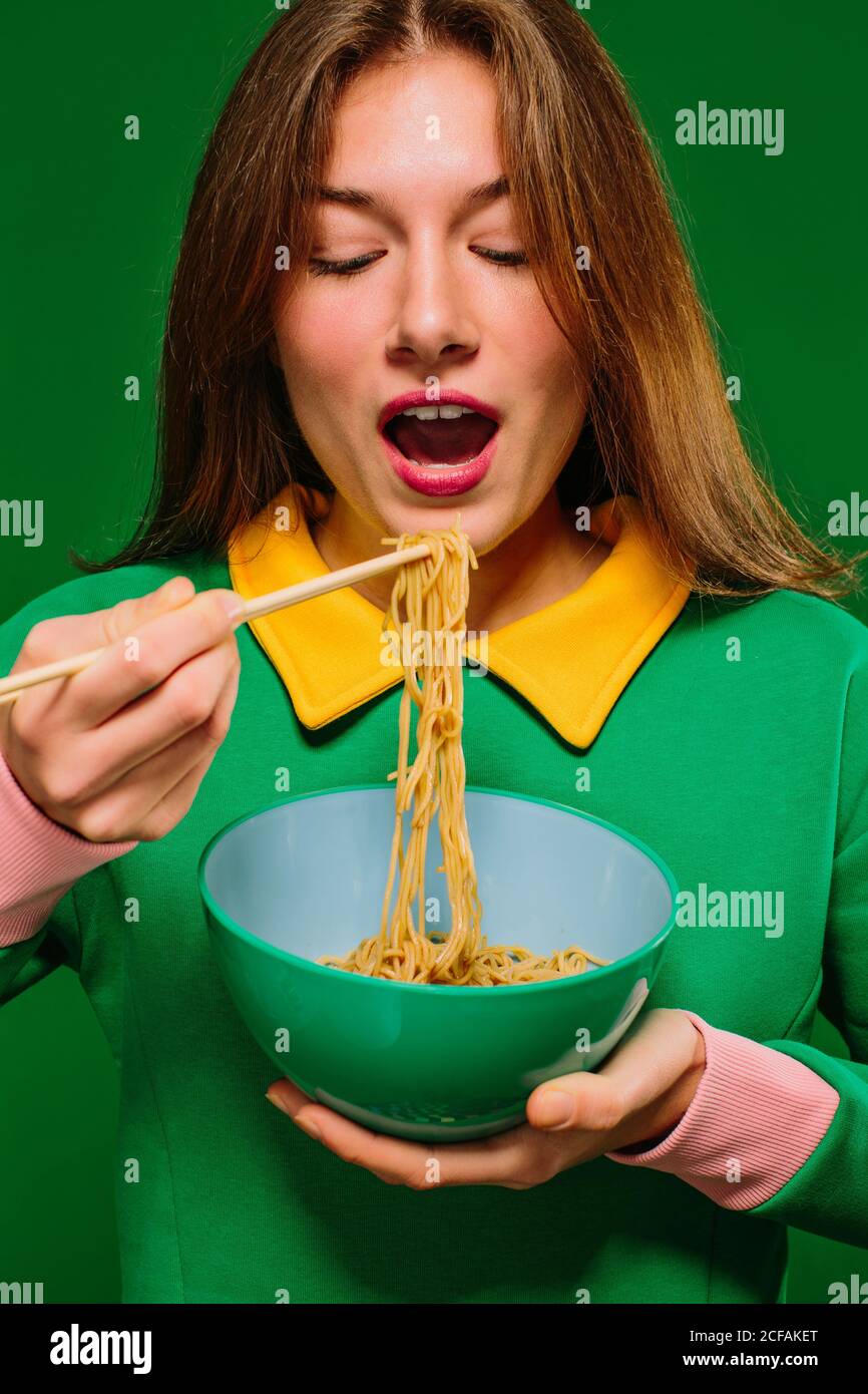 Giovane donna positiva in camicia verde con occhi chiusi mentre apertura bocca per mangiare noodle istantaneo yummy con chopsticks on sfondo verde Foto Stock