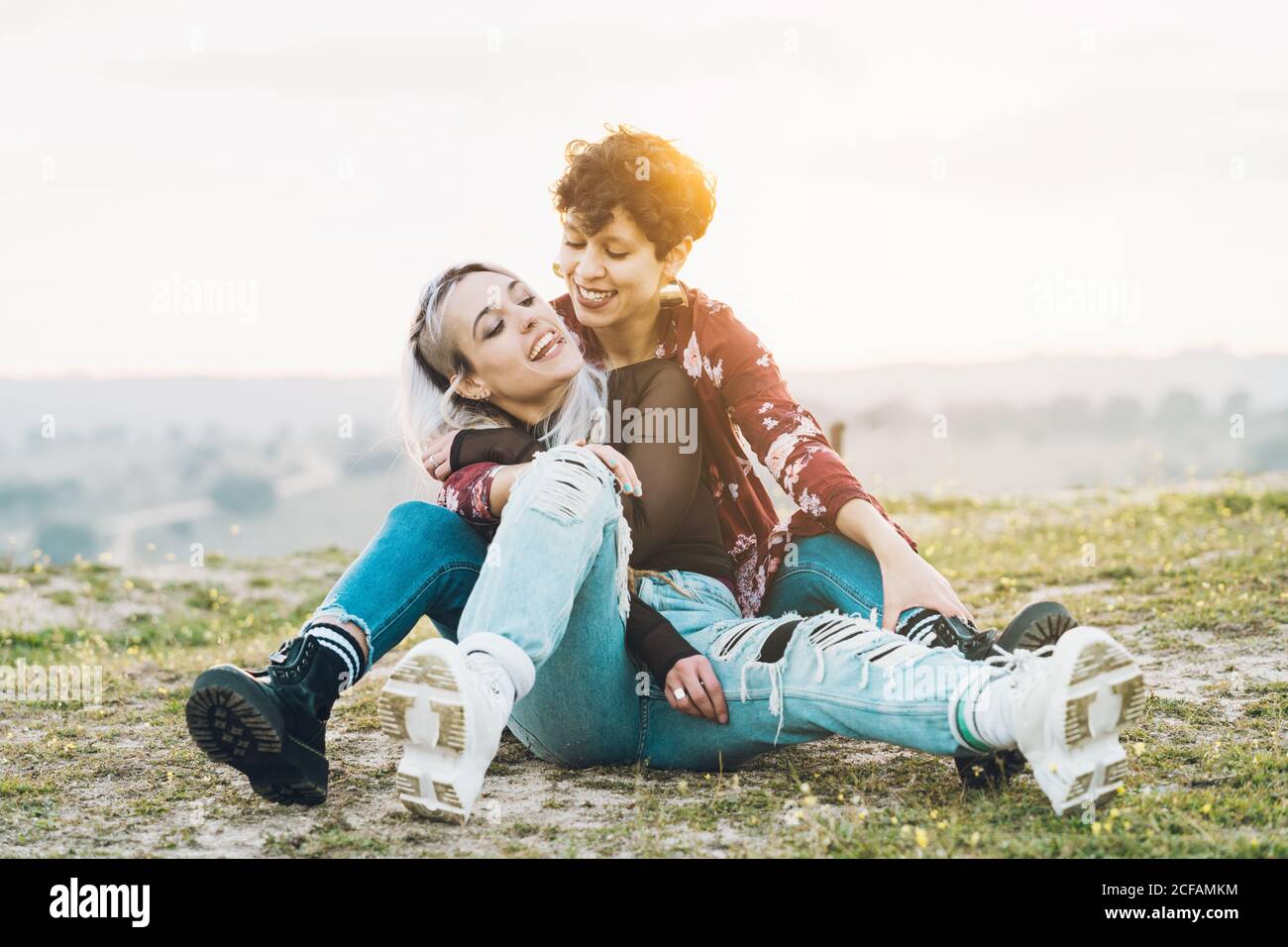 Ridendo le amiche strette in abiti casual eleganti seduti sul verde erba in abbraccio con cielo su sfondo Foto Stock