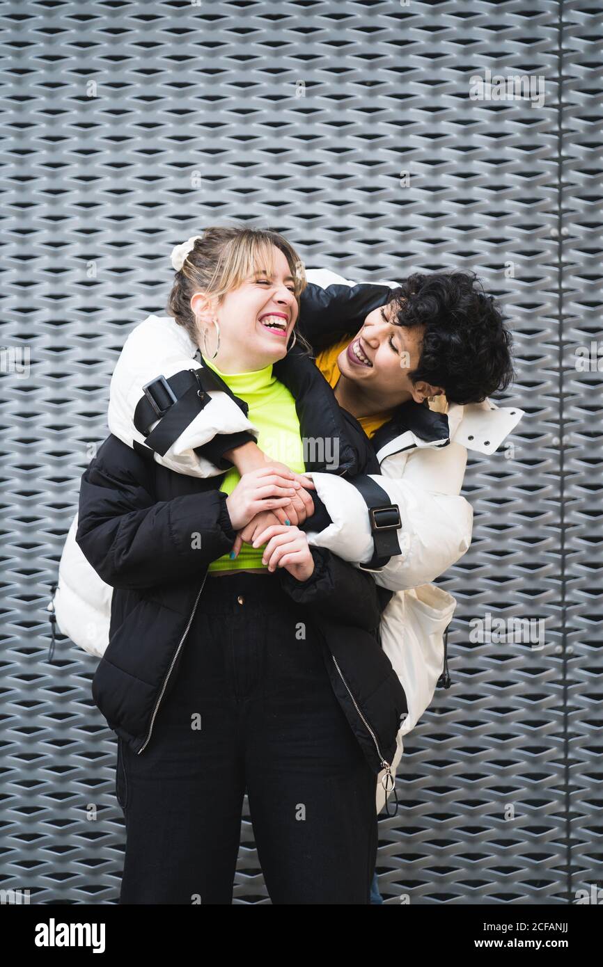 Contenuto allegro multietnico amici femminili in elegante abbigliamento caldo in piedi in abbraccio e sorridente con un edificio moderno sullo sfondo Foto Stock