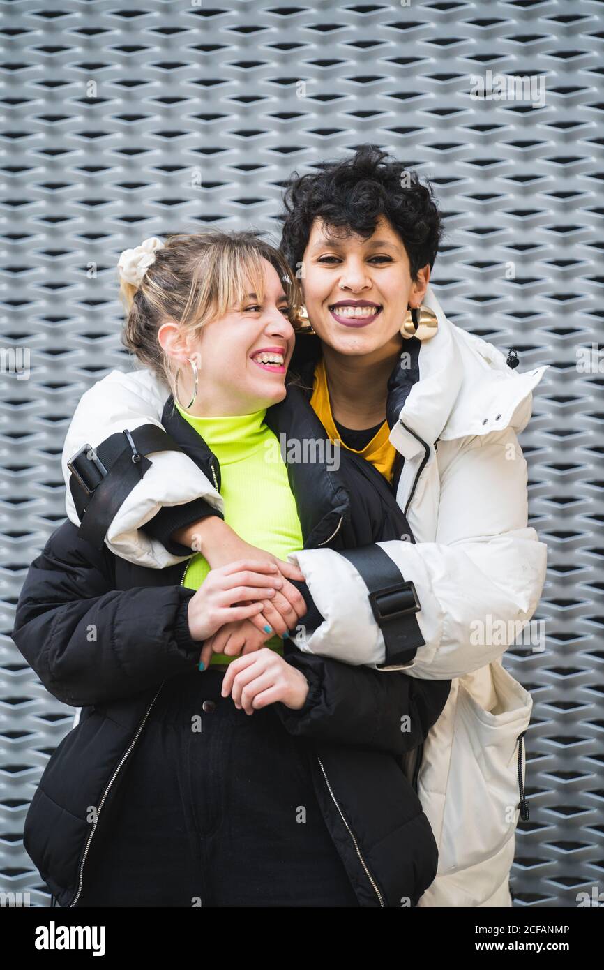 Contenuto allegro multietnico amici femminili in elegante abbigliamento caldo in piedi in abbraccio e sorridente con un edificio moderno sullo sfondo Foto Stock