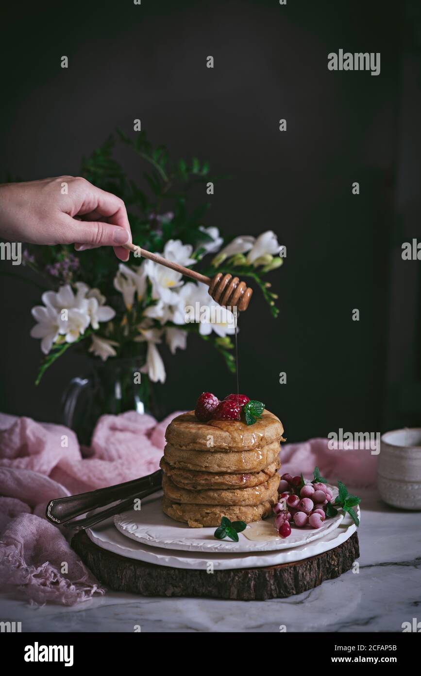 Pila di frittelle di lamponi con frutti di bosco freschi e miele gocciolante su piastra in porcellana sul piano portapaziente in marmo, testo da inserire nel contenitore con fiori su sfondo scuro Foto Stock