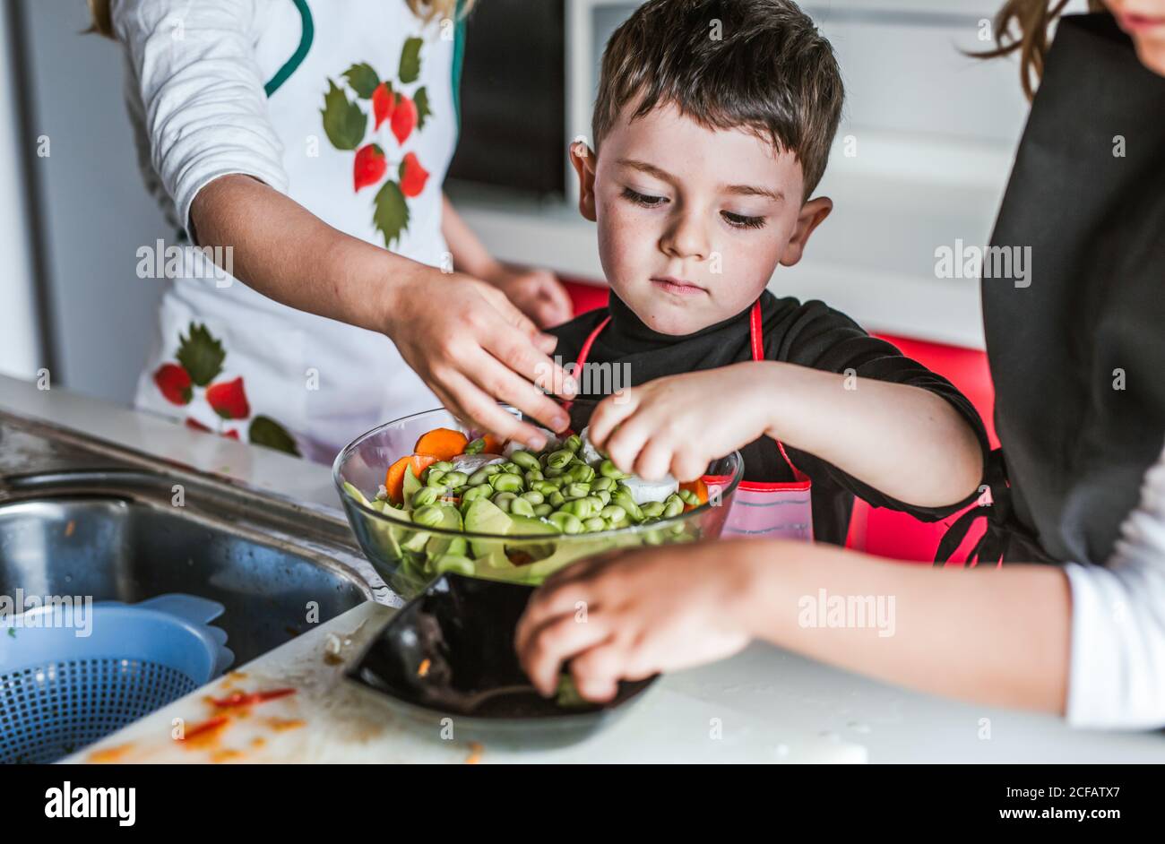 Bambine e ragazzo tagliando e peeling verdure mature mentre cucina sana insalata in cucina insieme Foto Stock