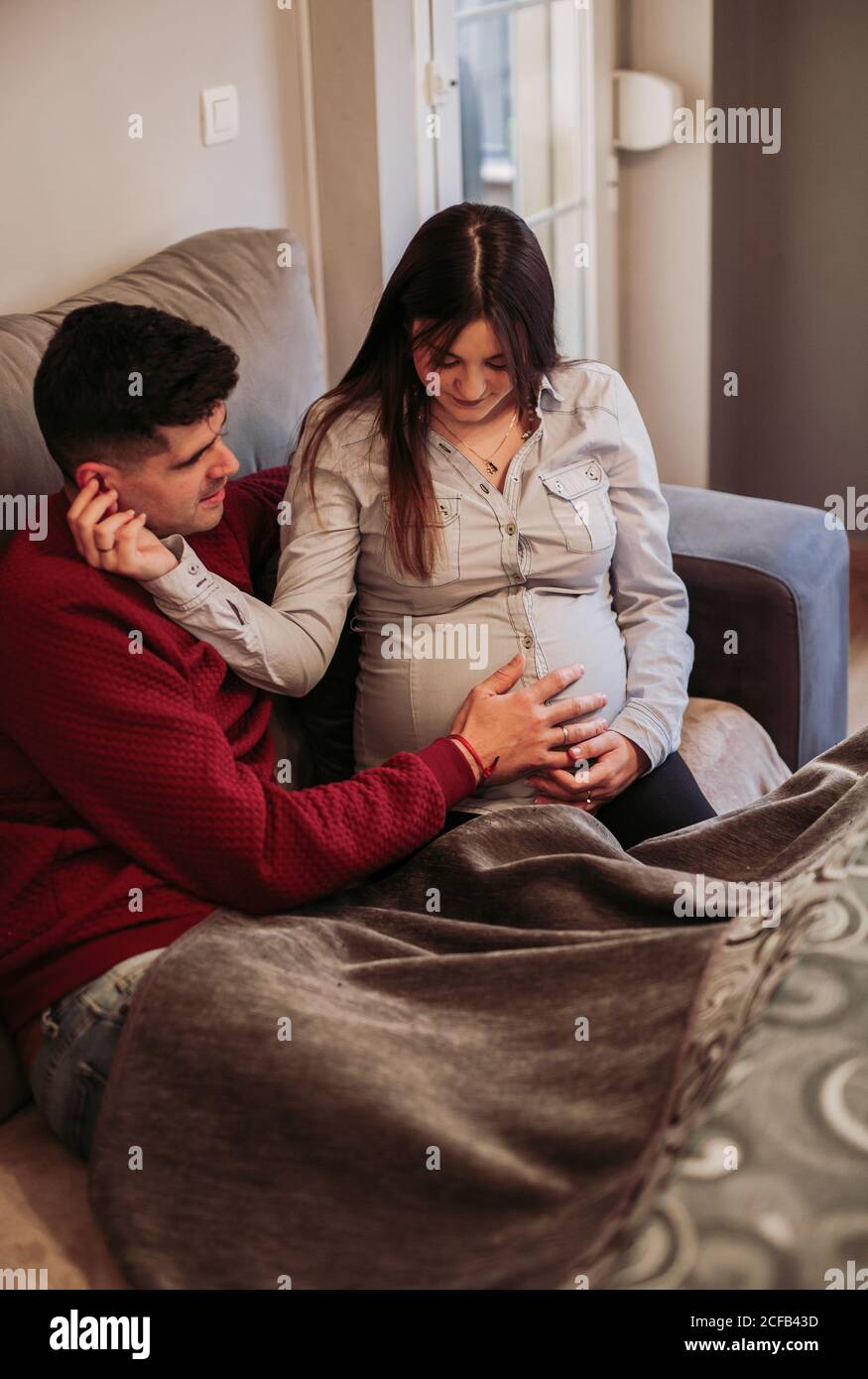 Giovane uomo in ponticello rosso seduto con la moglie incinta dentro blusa beige sul divano e tocchi il bambino mentre si spende insieme giorno d'inverno a casa Foto Stock