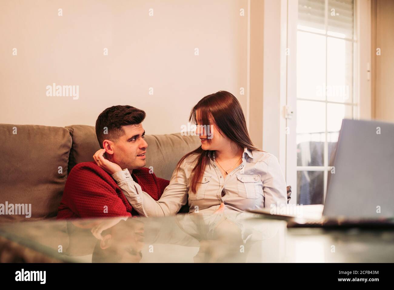 Giovane uomo in ponticello rosso seduto con la moglie incinta dentro blusa beige sul divano e tocchi il bambino mentre si spende insieme nella giornata invernale del computer a casa Foto Stock