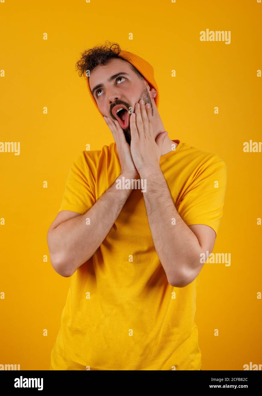 Uomo trendy bearded in vestito giallo che fa gesturing divertente del viso sorpresa guardando in piedi su sfondo giallo luminoso Foto Stock
