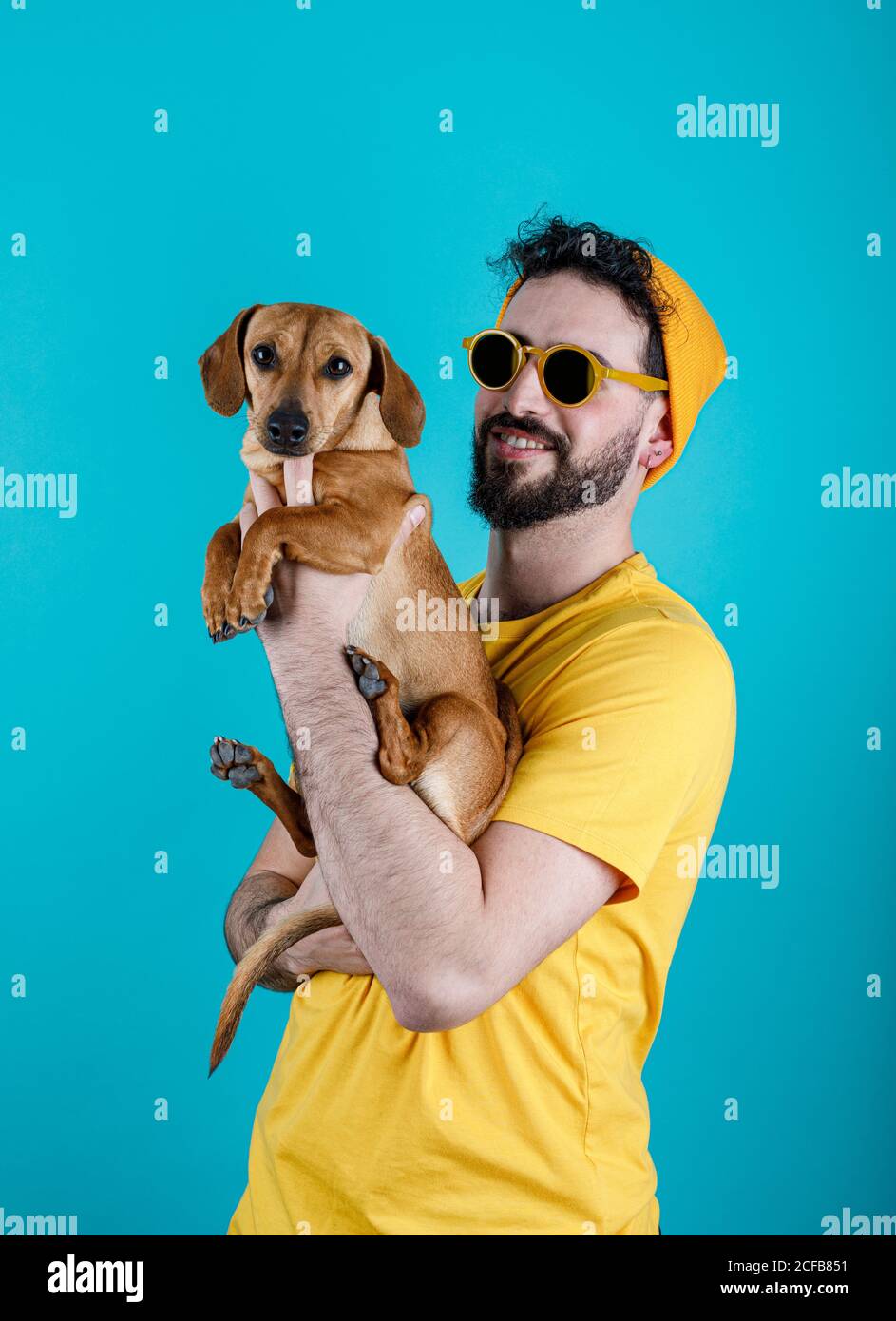 Allegro ragazzo sopportato in abito giallo alla moda sorridente e trasportante Carino cane Dachshund su sfondo turchese Foto Stock