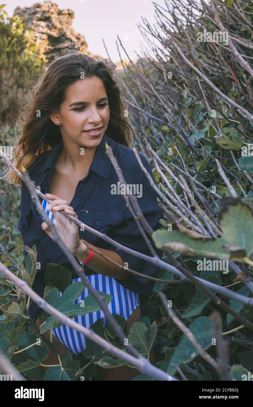 Elegante ragazza capelli ricci turista in camicia verde scuro e. bodysuit guardando via mentre camminando sulla foresta con fico albero sullo sfondo Foto Stock