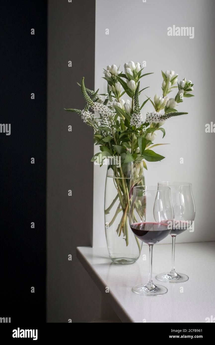 Un bouquet di veronica bianca con campane bianche in un vaso alto e stretto su un tavolo grigio su sfondo bianco. Vino rosso in alti bicchieri di vetro. Foto Stock