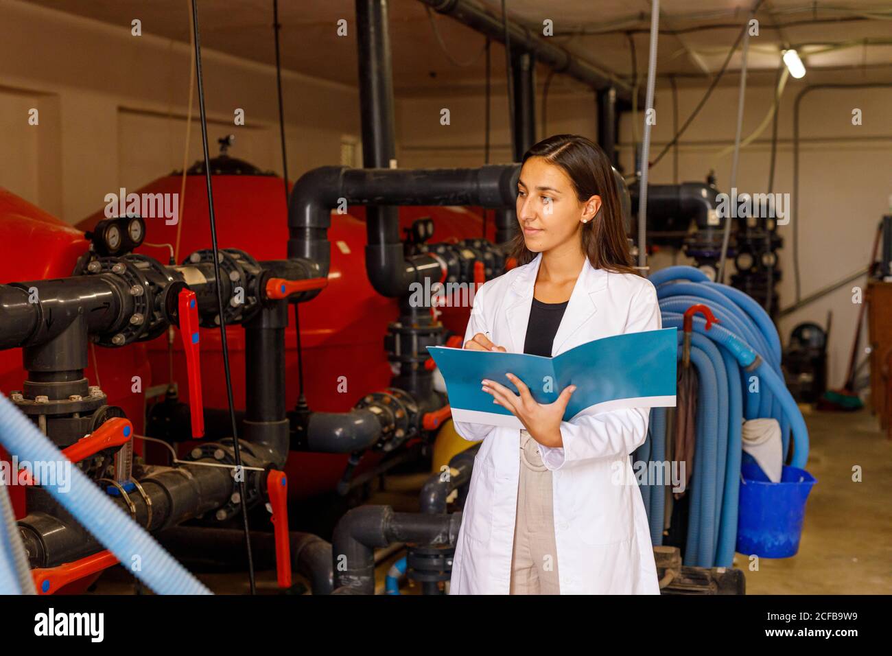 Giovane donna in laboratorio di controllo del rivestimento sistema di tubazioni e scrittura nel giornale durante l'ispezione di sicurezza in impianti industriali Foto Stock
