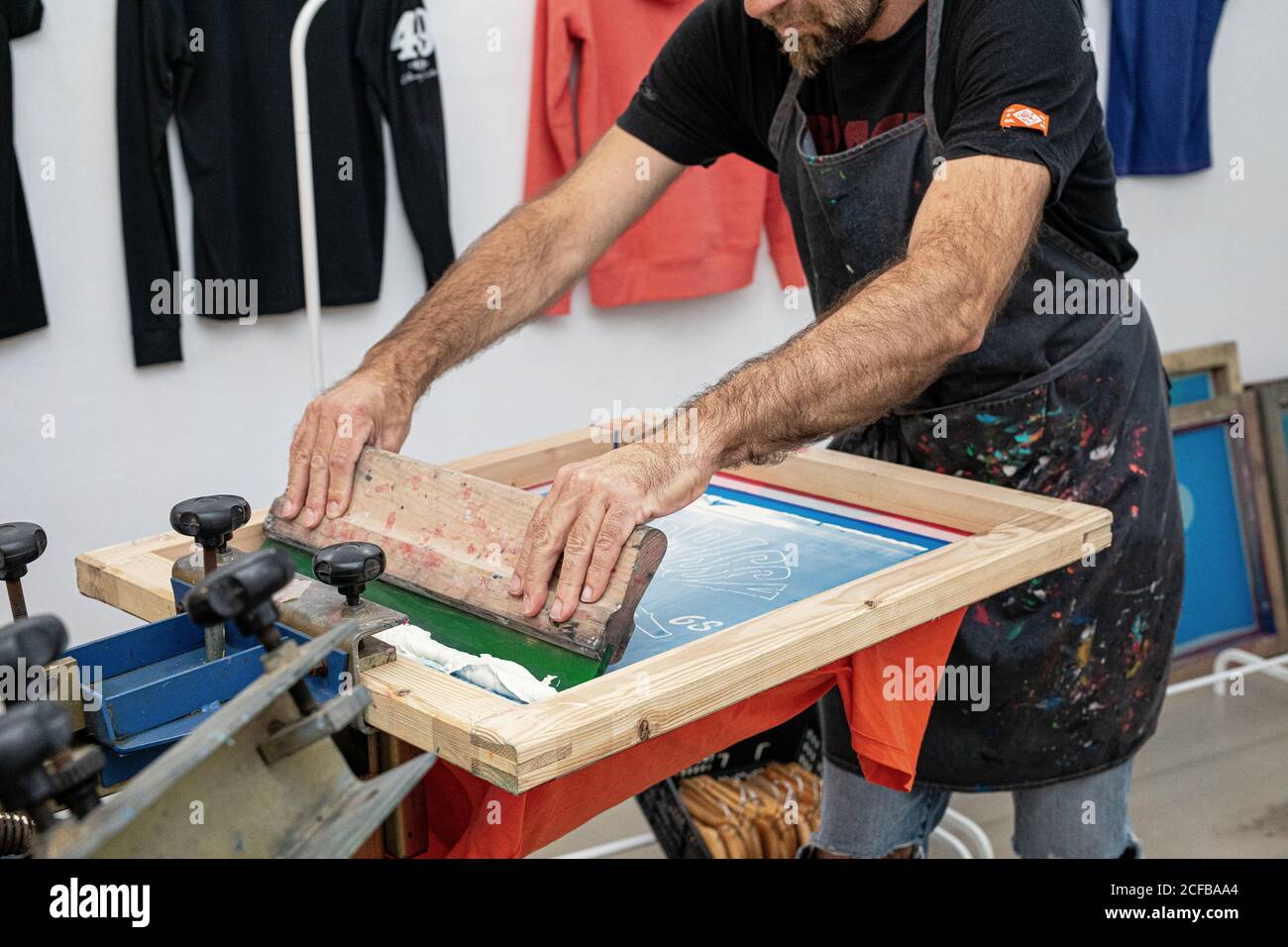 Crop uomo in grembiule sporco lavorando con serigrafia durante la creazione stampa su t-shirt in officina Foto Stock