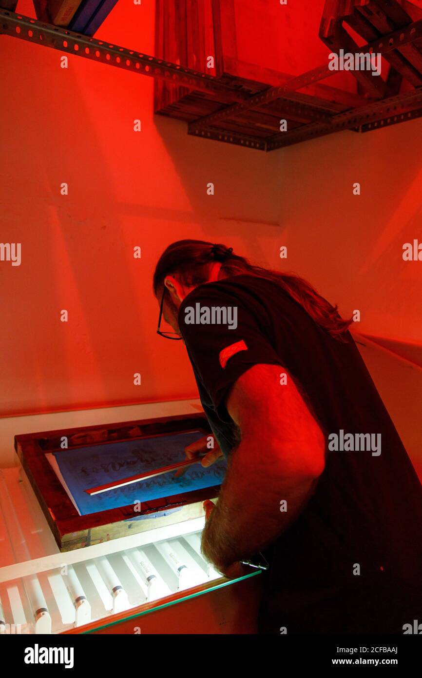 Artista maschile focalizzato con capelli lunghi asciugando stampa schermo rosso sala illuminata dell'officina Foto Stock