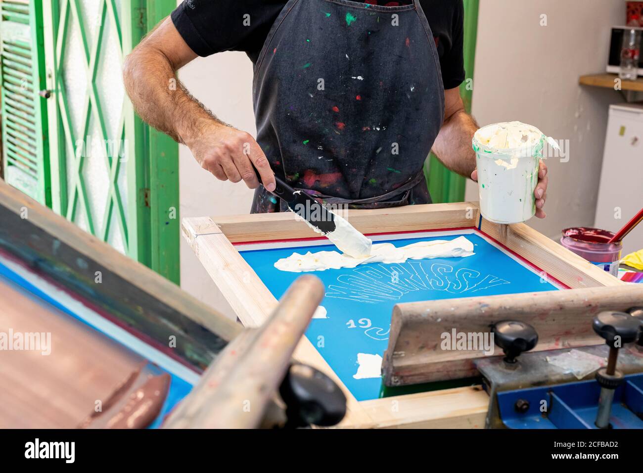 Crop uomo in grembiule sporco lavorando con serigrafia durante la creazione stampa su t-shirt in officina Foto Stock