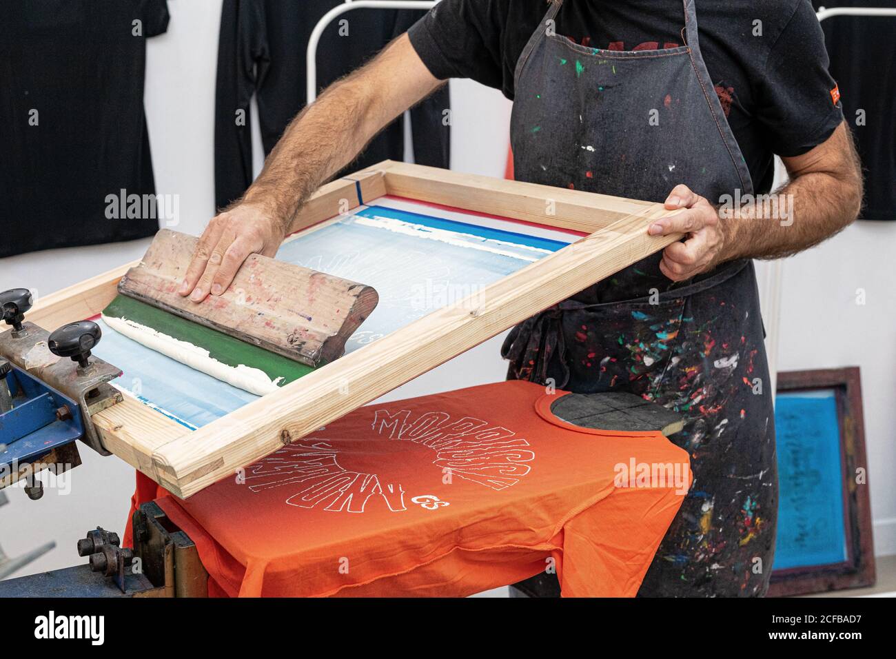 Crop uomo in grembiule sporco lavorando con serigrafia durante la creazione stampa su t-shirt in officina Foto Stock