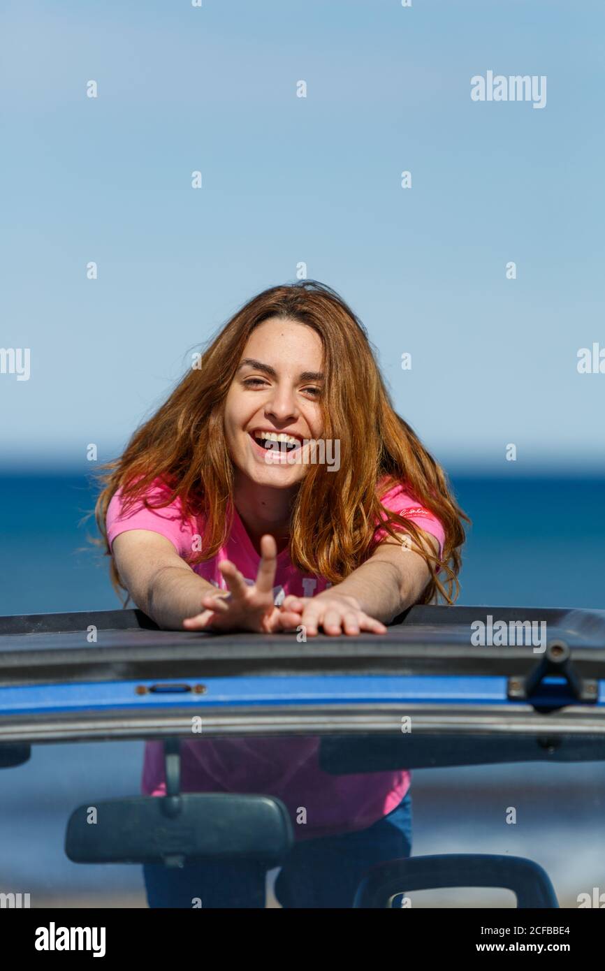 Attraente donna dai capelli lunghi che ride e guarda la macchina fotografica che posa sul tetto blu dell'automobile in giorno sereno luminoso Foto Stock