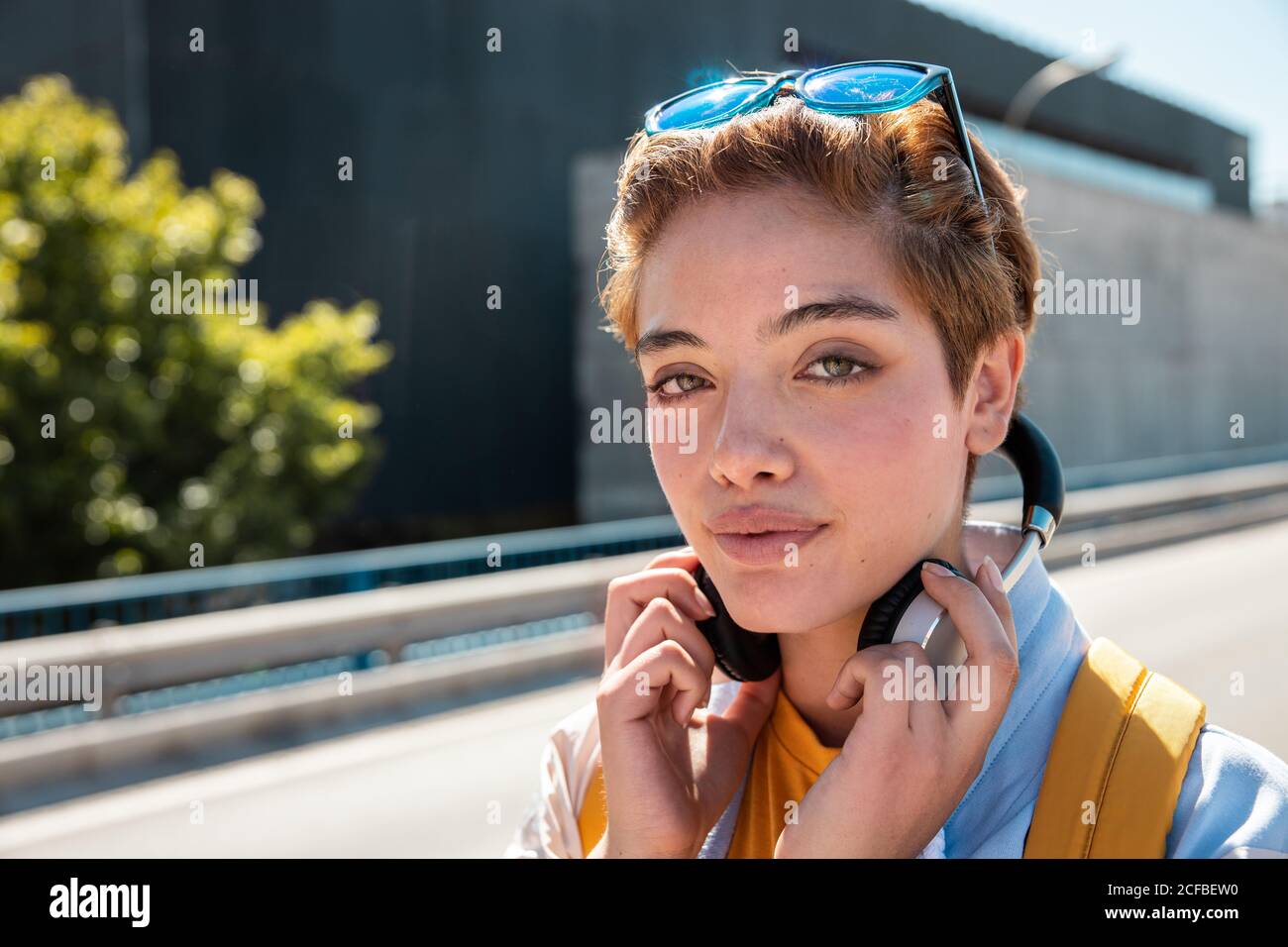 Donna millennial elegante e snello, magnifica con occhi verdi incredibili e un sorriso tondo in abiti bianchi e gialli con cuffie wireless e occhiali da sole che guardano la fotocamera contro le strade sfocate nelle giornate di sole Foto Stock