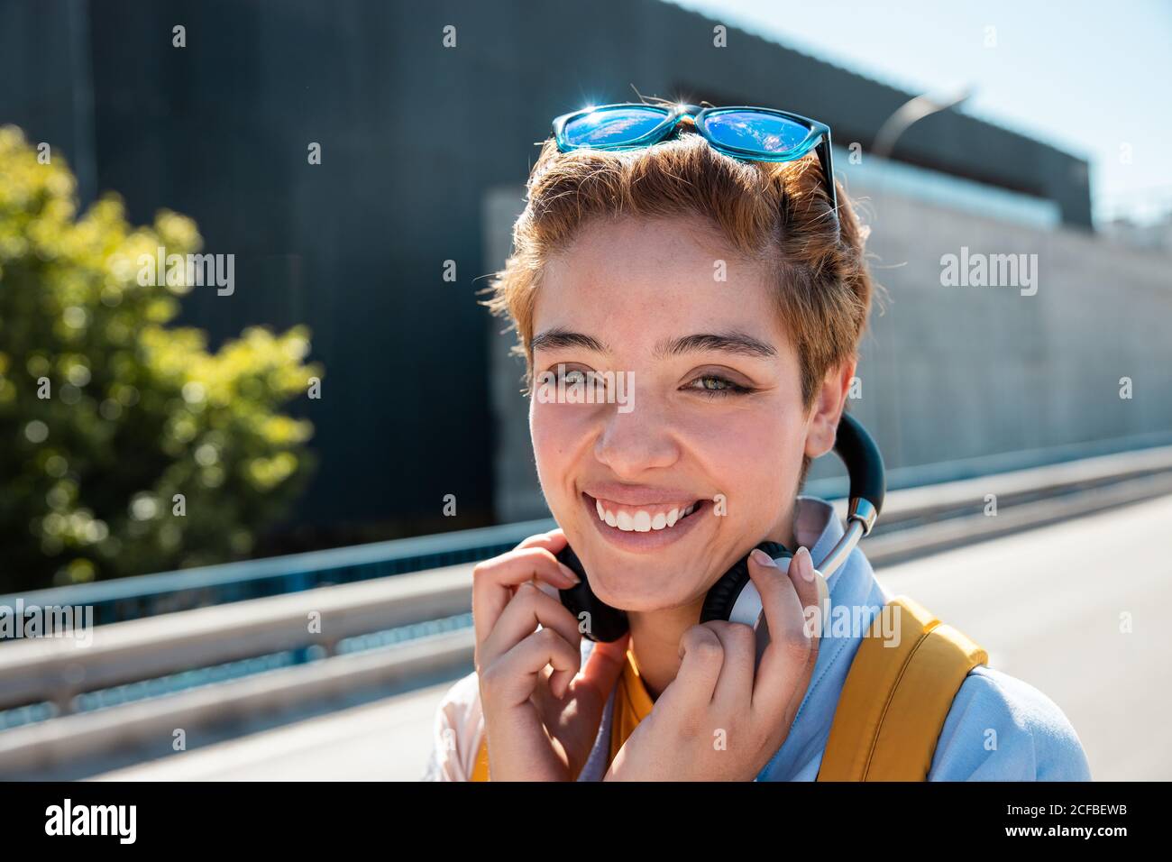 Donna millennial elegante e snello, magnifica con occhi verdi incredibili e un sorriso tondo in abiti bianchi e gialli con cuffie wireless e occhiali da sole che guardano la fotocamera contro le strade sfocate nelle giornate di sole Foto Stock