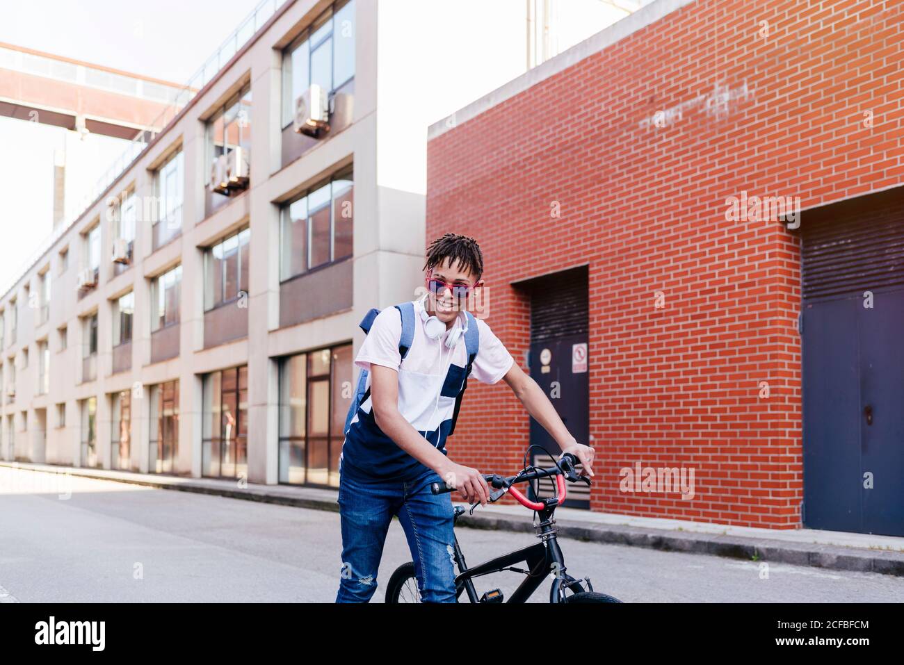 Giovane uomo nero sorridente di successo con capelli afro in casual outfit e occhiali da sole eleganti in piedi con bicicletta sulla strada Foto Stock