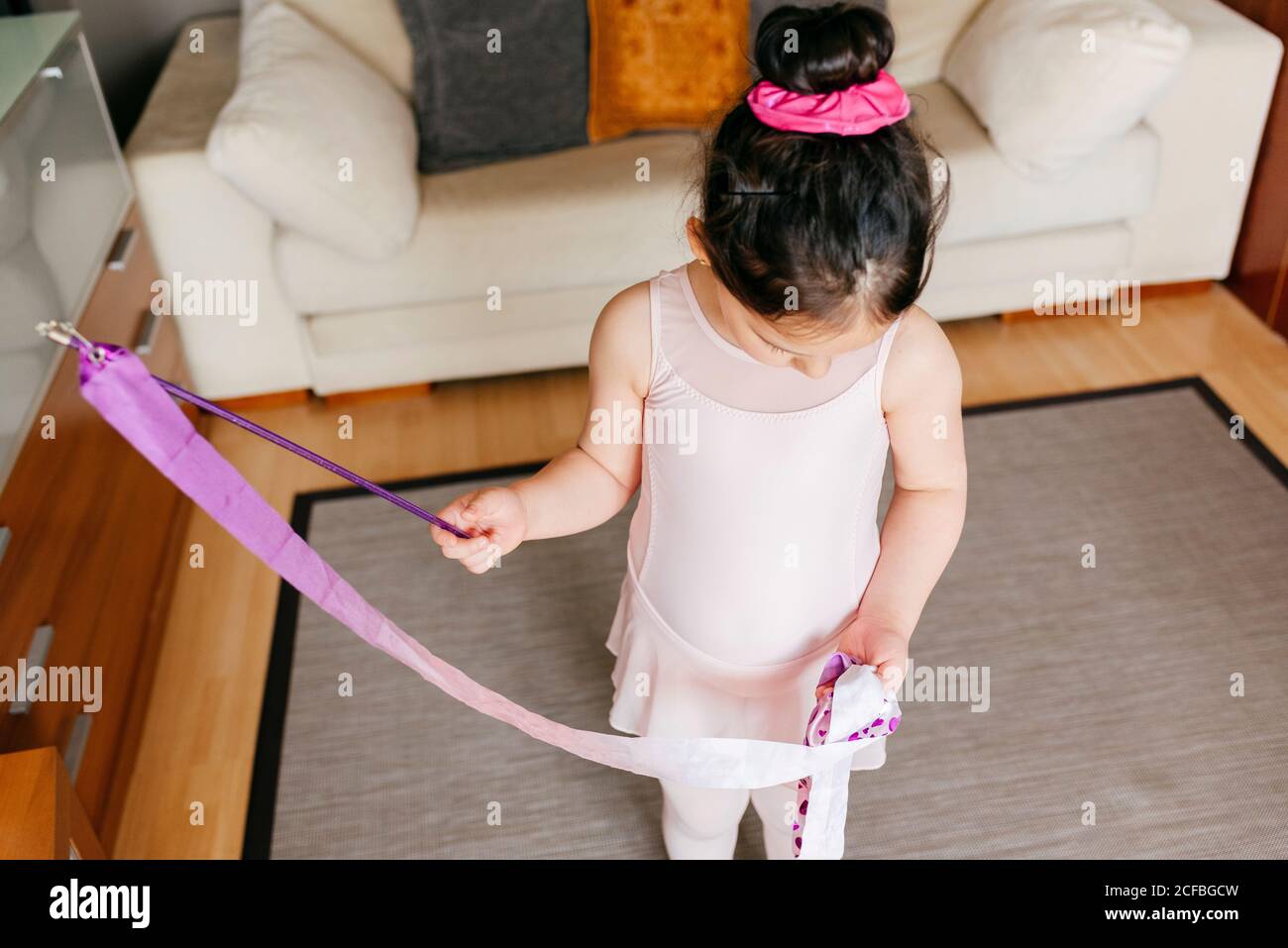 Dall'alto ha messo a fuoco la bambina carina in leotard e tights filatura del nastro durante l'allenamento di ginnastica ritmica in una vita accogliente camera a casa Foto Stock