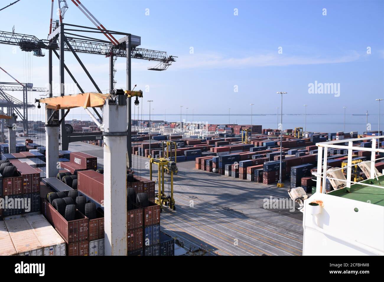 Vista dal ponte della nave mercantile sotto carico su un molo di container terminal coperto di container pronti per essere caricati sotto il cielo blu. Foto Stock