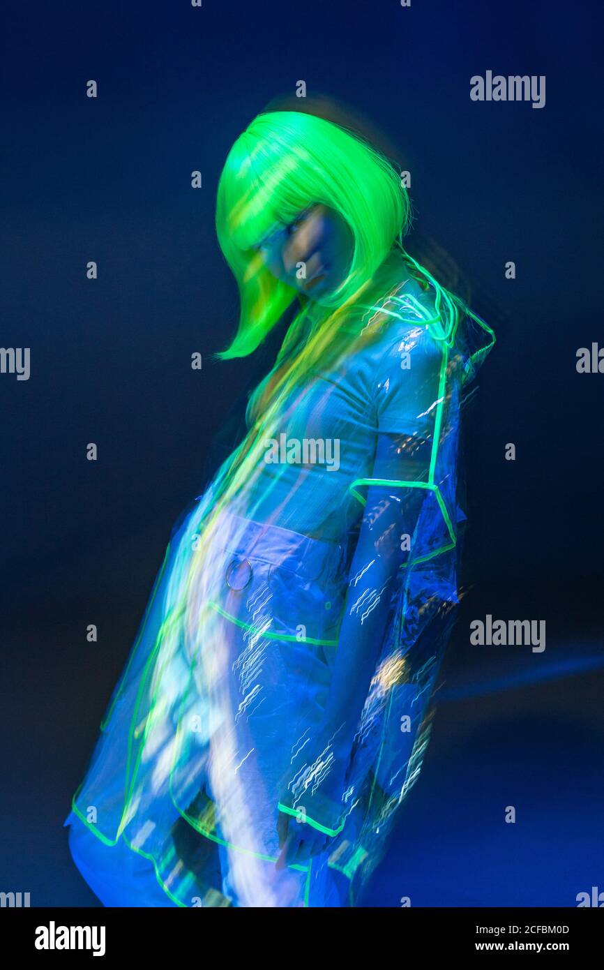 Giovane donna asiatica in parrucca gialla e materiale plastico trasparente che posa in luce fluorescente Foto Stock