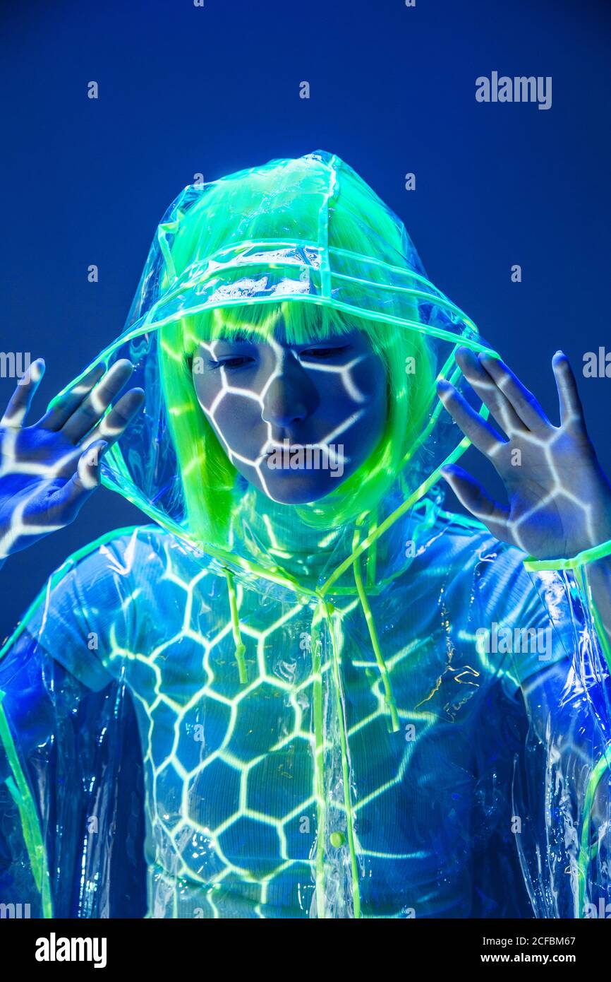 Giovane donna asiatica in parrucca gialla e materiale plastico trasparente che posa in luce fluorescente con mani sollevate Foto Stock