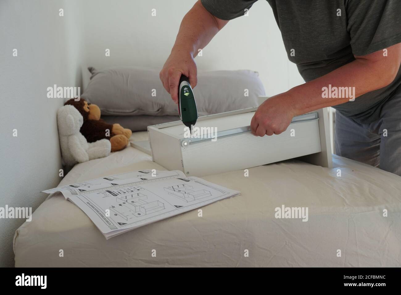 Un uomo che monta un cassetto di una scrivania per computer per suo figlio utilizzando un cacciavite a batteria. Sta utilizzando uno strumento e le istruzioni per completare il progetto. Foto Stock