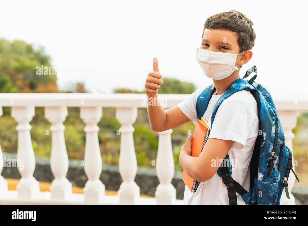 Ritratto di di bambino di scuola che indossa maschera protettiva per l'inquinamento o virus e mostrando pollici in su. Coronavirus e concetto scolastico Foto Stock