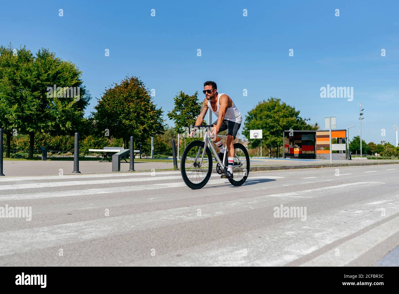 Uomo atletico in occhiali da sole con camicia bianca e pantaloncini neri pedalando sulla strada della città con alberi verdi sulla strada il giorno d'estate con cielo blu Foto Stock