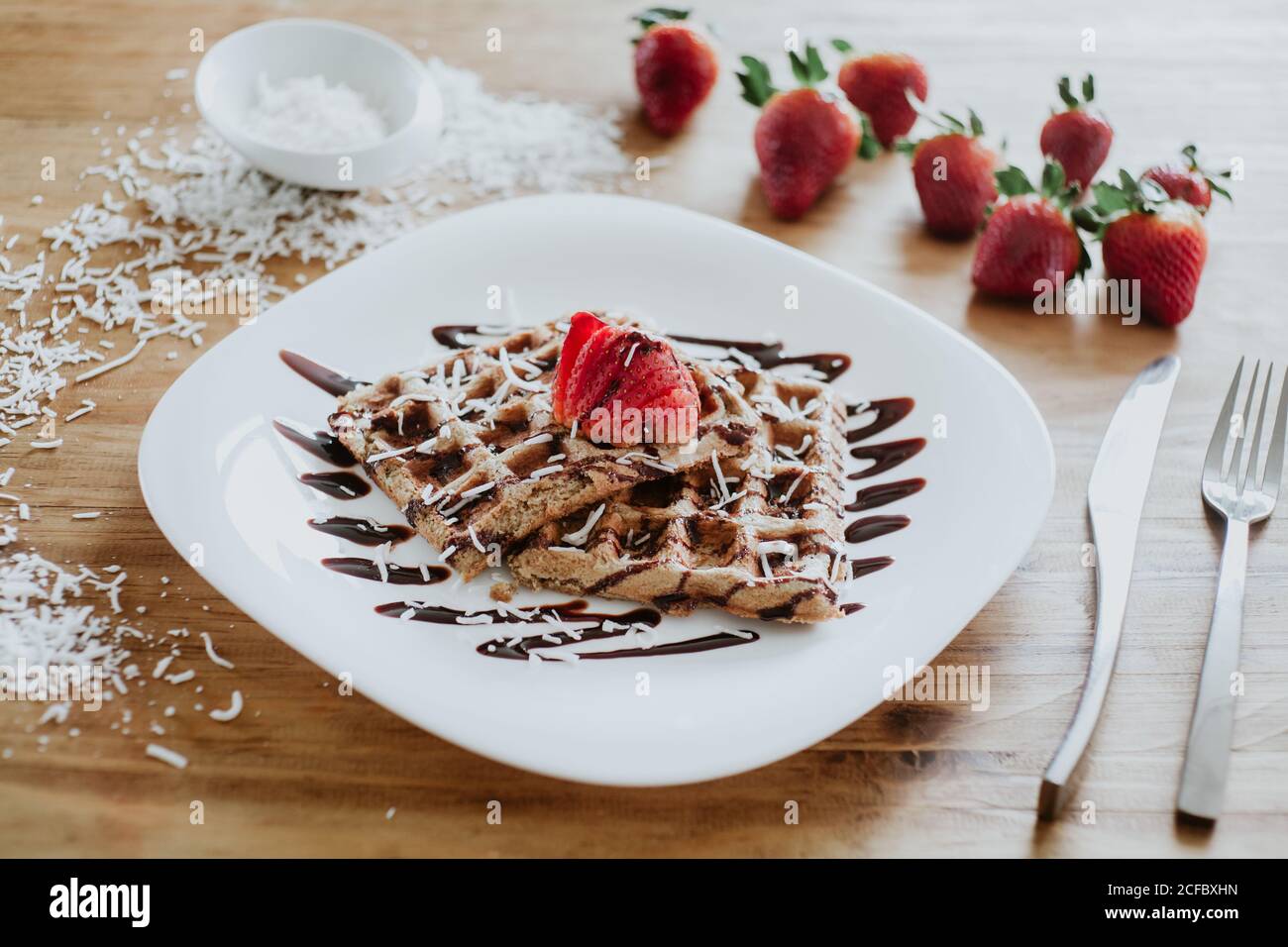 Cialde dolci e fragole servite sul piatto con yummy sciroppo di cioccolato e fiocchi di cocco su tavola di legno vicino argenteria Foto Stock