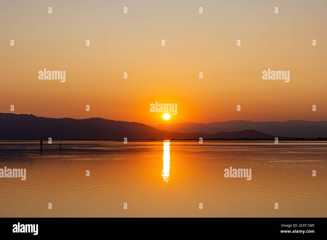 Il sole arancione scende dietro le colline scure riflettendosi in pace acqua leggermente increspata creando romantici condimenti Foto Stock