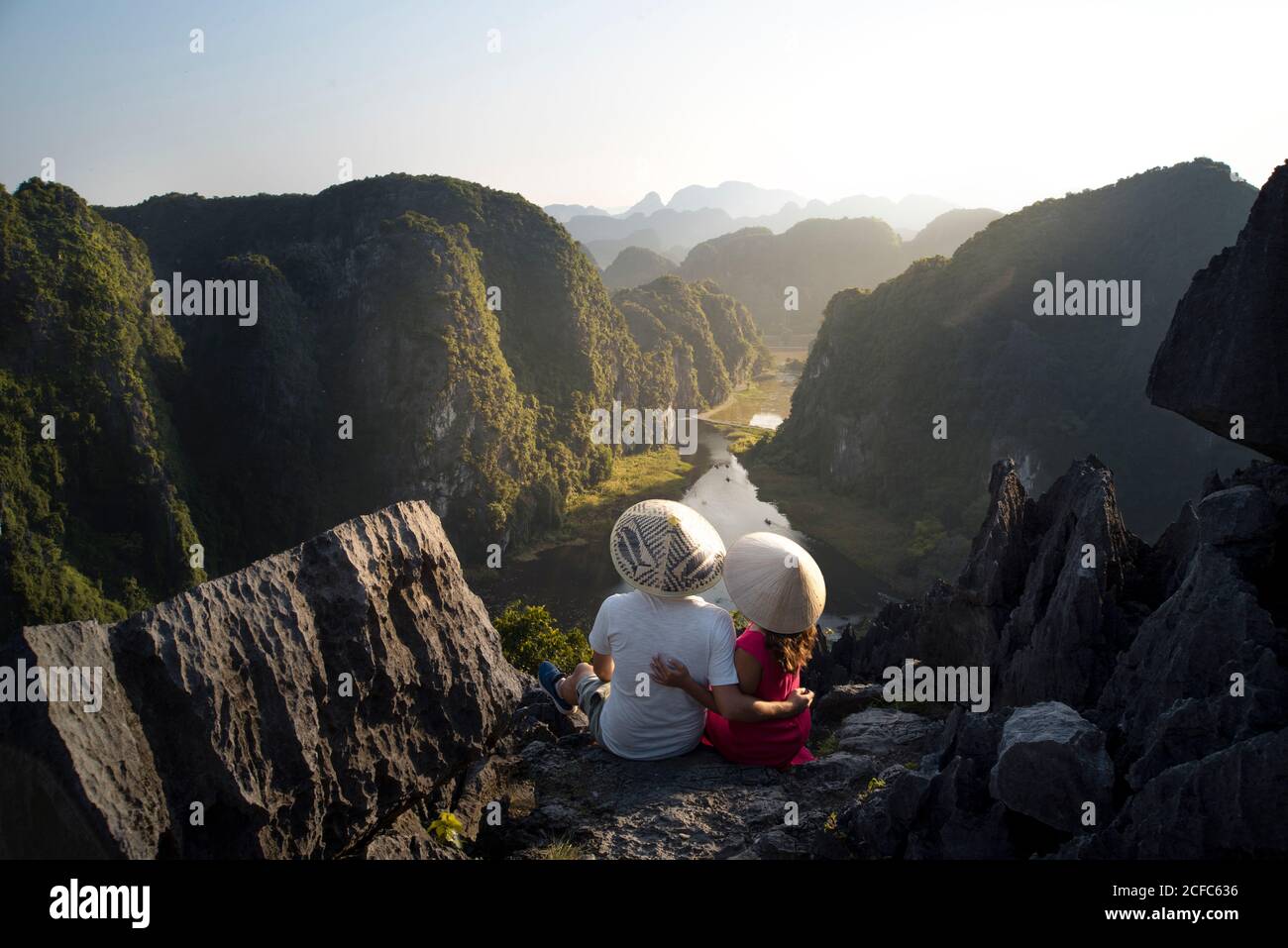 Da sopra vista posteriore di coppia di turisti in conico I cappelli abbracciano mentre si siedono sull'alta cima rocciosa di Mua caverna e ammirare la magnifica valle tropicale in creste Foto Stock