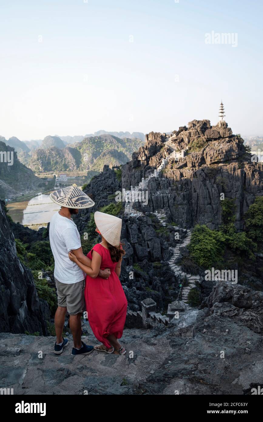 Dall'alto vista posteriore di coppia di turisti in tradizionale cappelli conici in piedi su una collina rocciosa e godendo di una vista maestosa Di Mua grotta mentre coccolarsi e guardare l'un l'altro Foto Stock