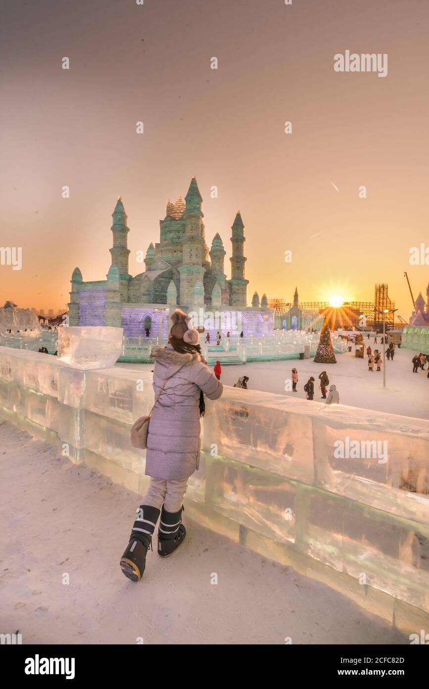 Vista posteriore di donna anonima in outwear caldo che pende sopra barriera di ghiaccio e ammirare sculture di neve in serata durante l'inverno festival Foto Stock