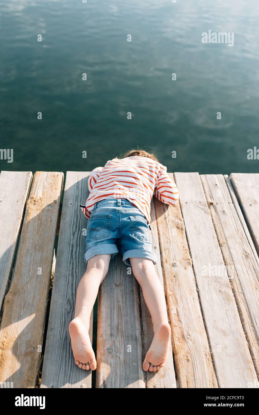 Dall'alto di bambino agghiacciante mentendo con faccia in giù sopra molo in legno al sole con acqua tranquilla sotto Foto Stock