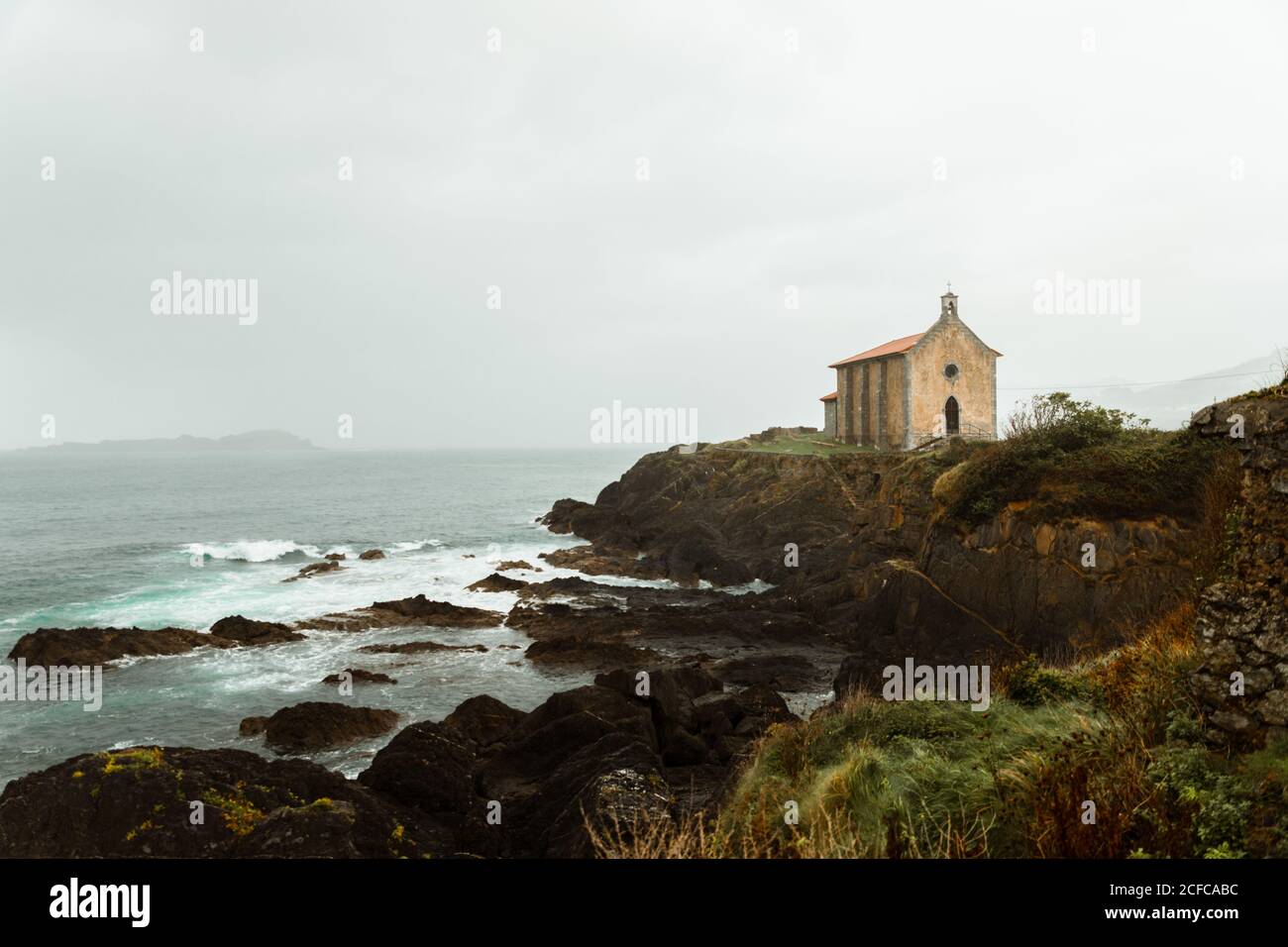 Oceano con grandi pietre vicino collina con antico edificio e. montagna lontano sotto il cielo bianco Foto Stock