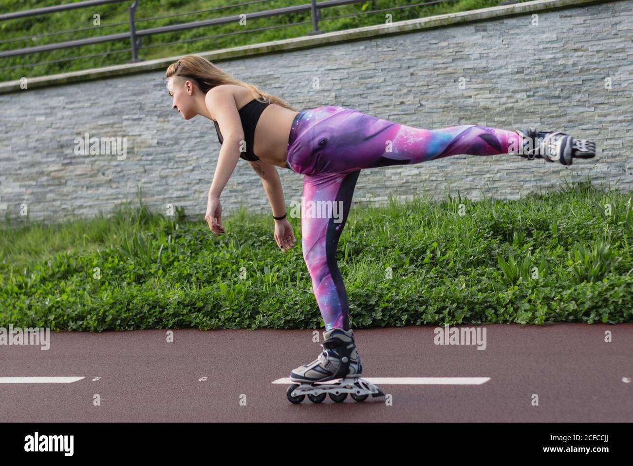 Vista laterale della giovane femmina in abbigliamento sportivo e lame a rulli equilibrio sulla gamba mentre divertente durante il fine settimana Foto Stock