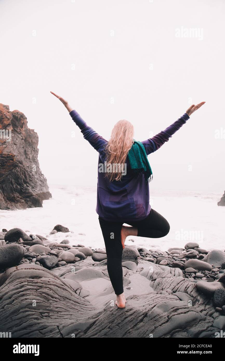 Giovane Donna con mani alzate meditando e stando in piedi sulle rocce vicino alla costa del mare Foto Stock