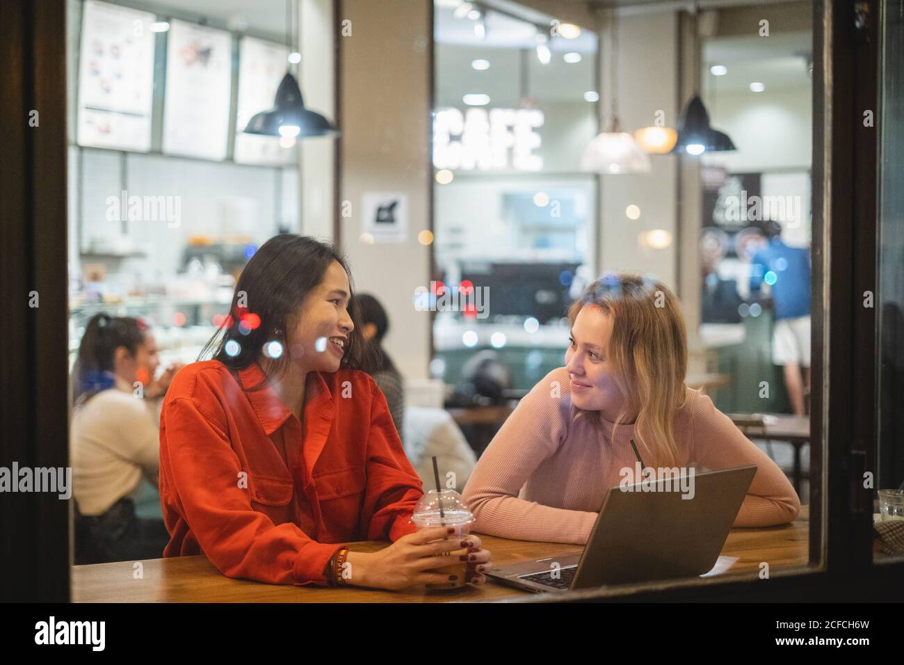 Giovani donne multirazziali sorridenti e che parlano tra loro seduto al tavolo in un'accogliente caffetteria che lavora sul computer Foto Stock