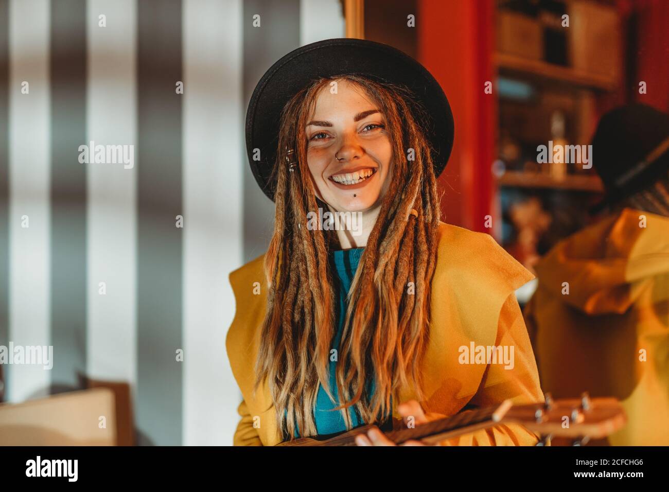 Giovane donna allegra ed elegante con i dreadlock che indossano il cappotto  giallo e. cappello nero seduto sul vecchio tavolo di legno di ritorno a  specchio E suonare la chitarra hawaiana ukulele