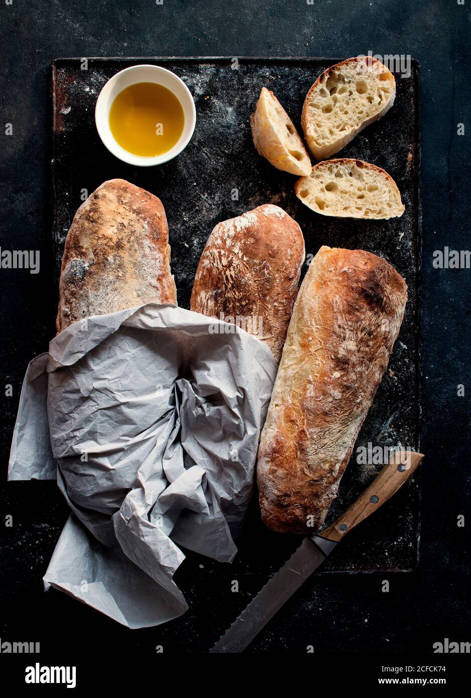 Pane ciabatta su tavola rustica vicino all'olio d'oliva e coltello con fette di pane su sfondo scuro Foto Stock