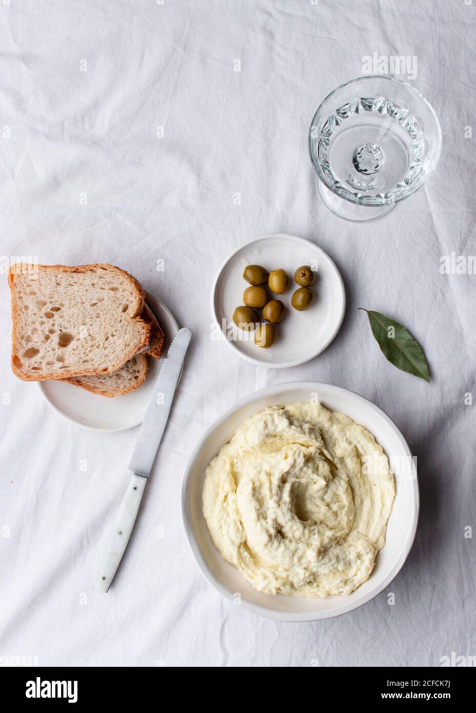 Da sopra ciotola con brandade di merluzzo posto vicino alle olive, fette di pane e foglie di alloro su un tavolo da cucina sfondo bianco Foto Stock