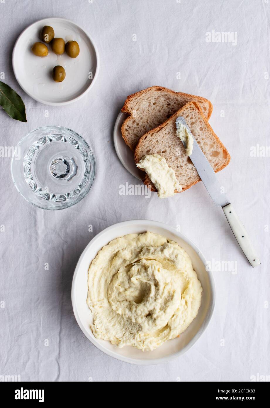 Da sopra ciotola con brandade di merluzzo posto vicino alle olive, fette di pane e foglie di alloro su un tavolo da cucina sfondo bianco Foto Stock