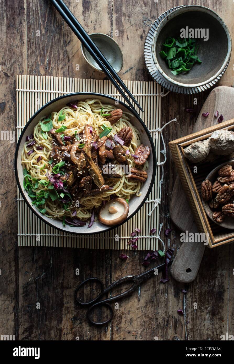 Da sopra ciotola con deliziosa pasta miso con piante d'uovo e funghi cinesi sul tappetino vicino a forbici, bastoncini, cipolla munga e zenzero sul tagliere Foto Stock