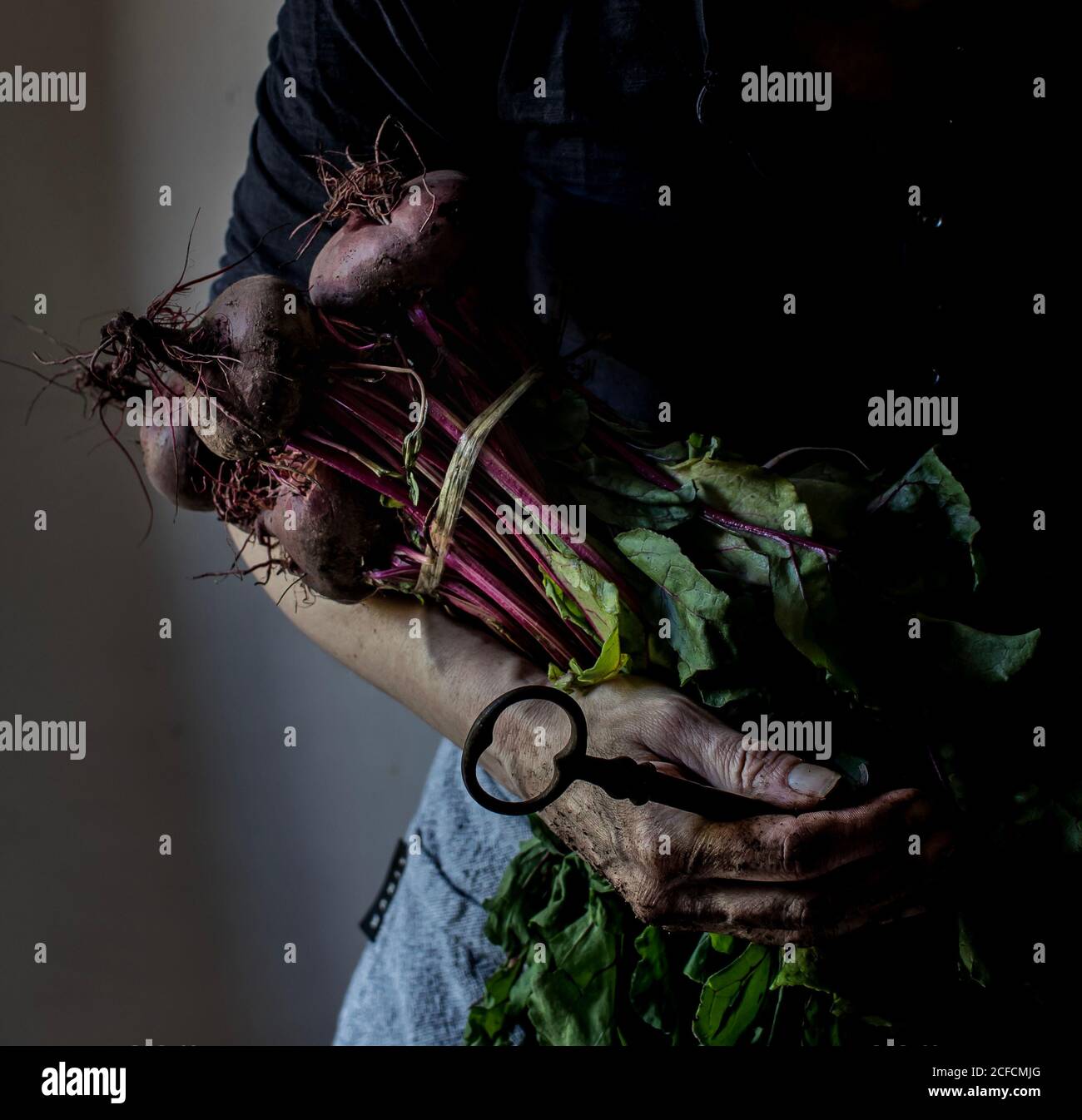 Crop Senior Woman che tiene mazzo di barbabietole fresche biologiche e grande chiave con mano sporca su sfondo grigio Foto Stock