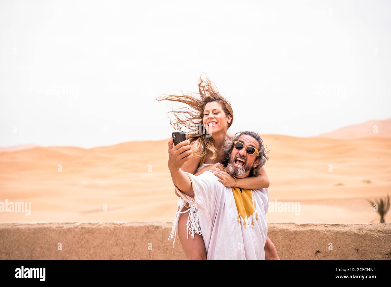 Uomo di mezza età con Piggyback donna che prende selfie in modo espressivo sulla terrazza contro il deserto sabbioso, Marocco Foto Stock