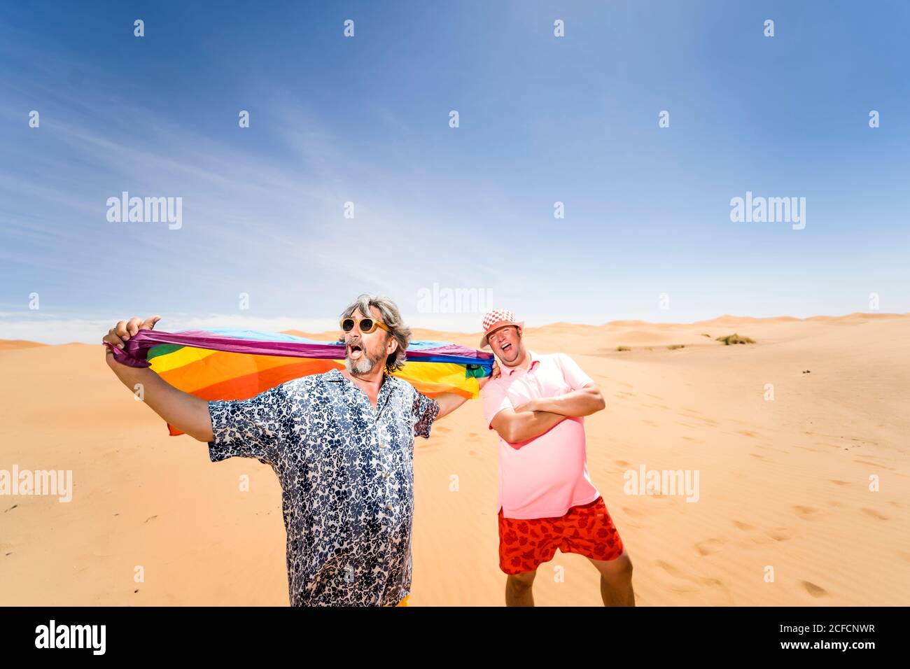 Felice sovrappeso uomini maturi che si divertono con la bandiera LGBT mentre si erge sullo sfondo di un arido deserto e di un cielo blu Foto Stock