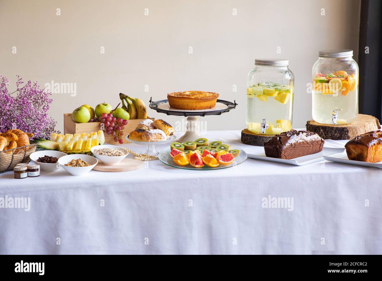 Vista laterale della cena festiva con gustosi prodotti da forno a base di frutta e banche eleganti di limonata su tavolo decorato con lilla fiori Foto Stock