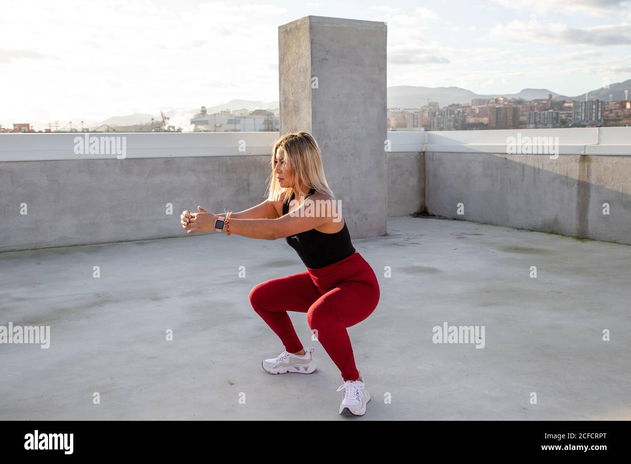 Donna concentrata con capelli biondi in abbigliamento attivo in posizione squat con braccia distese sul tetto dell'edificio Foto Stock