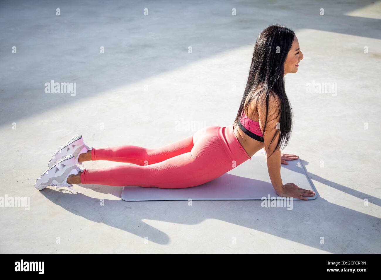 Dall'alto vista laterale della giovane donna dai capelli brunette in abbigliamento sportivo guardando lontano mentre si appoggia sulle mani diritte sdraiate sul tappeto sul tetto di cemento dell'edificio Foto Stock