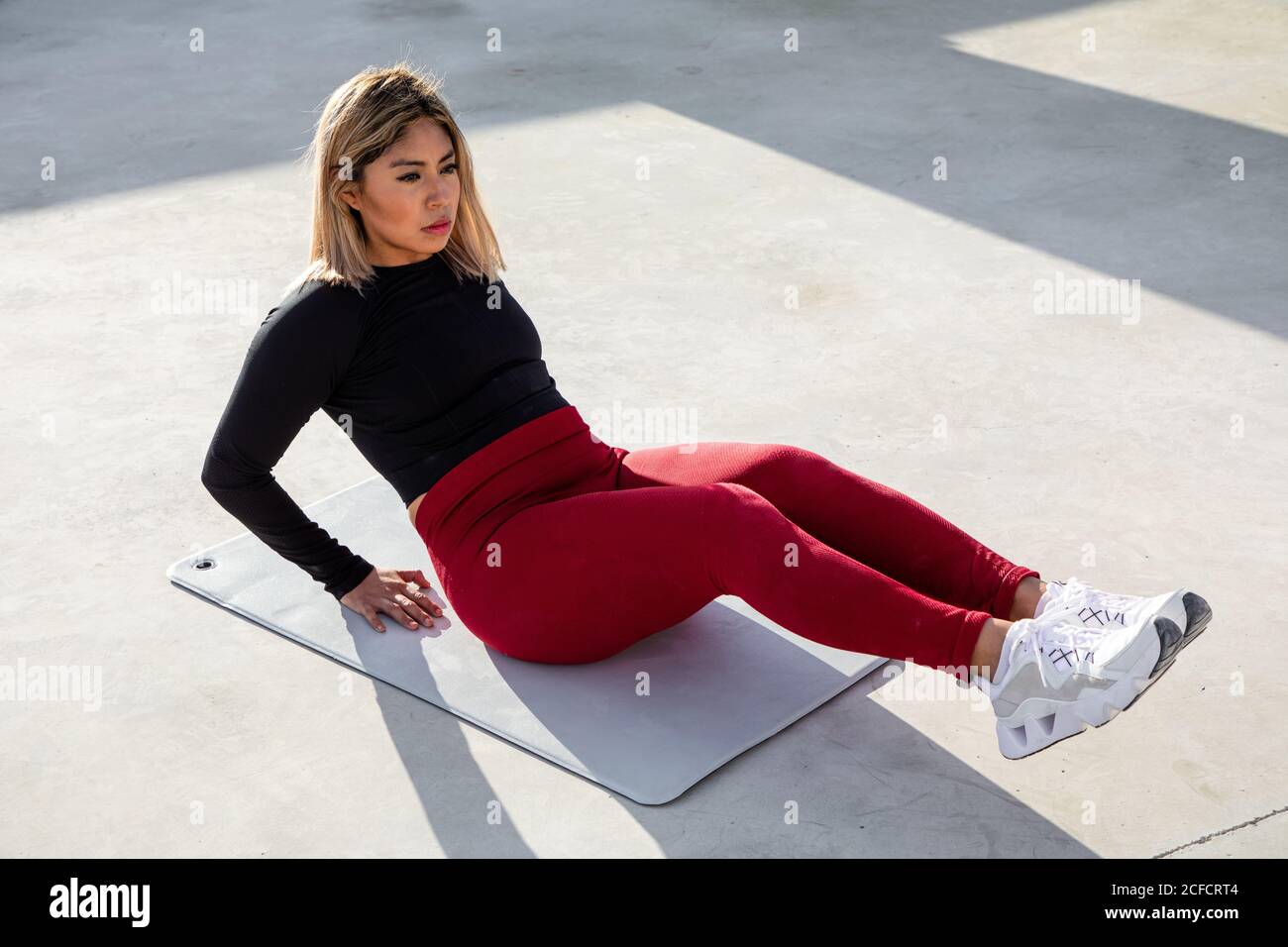 Dall'alto della donna con capelli biondi in abbigliamento sportivo seduta su tappetino con gambe piegate allenamento addome e guardando lontano sul tetto della costruzione Foto Stock